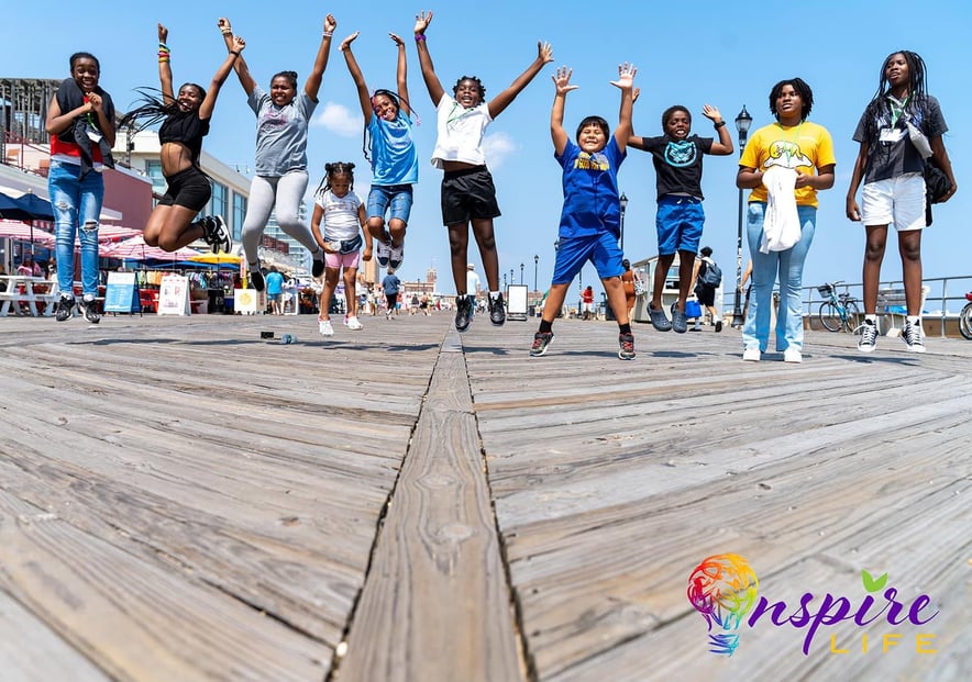 Children in a row jumping in the air on a boardwalk with bright blue skies. Inspire Life logo bottom right.